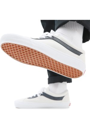 Weiße Sneakers mit schwarzen Streifen, getragen mit weißen Socken und schwarzen Hosen, ein Fuß angehoben, um die Waffelmuster-Sohle zu zeigen.