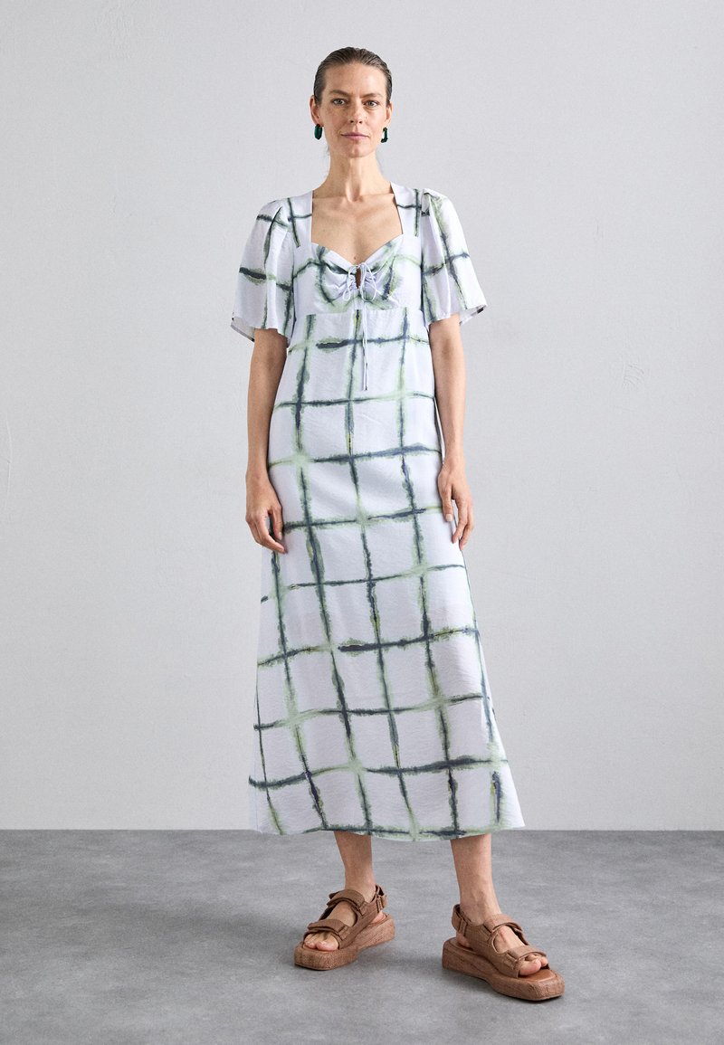 Maxi jurk van lichtblauwe stof met een groen en donkerblauw ruitpatroon, met pofmouwen en een strikdetail aan de voorkant. Gekoombineerd met bruine plateau-sandalen.