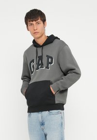 Hanorac gri din bumbac cu buzunar și manșete negre. Prezintă logo-ul "GAP" în text alb conturat. Buzunar tip kangaroo pe față, croială lejeră. Textură ușoară.