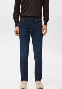 Mann trägt dunkelblaue Jeans mit geradem Bein, dunkelbraunen Pullover und beige Sneaker, steht vor einem einfarbigen Hintergrund.