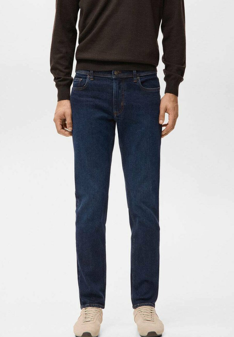 Mann trägt dunkelblaue Jeans mit geradem Bein, dunkelbraunen Pullover und beige Sneaker, steht vor einem einfarbigen Hintergrund.