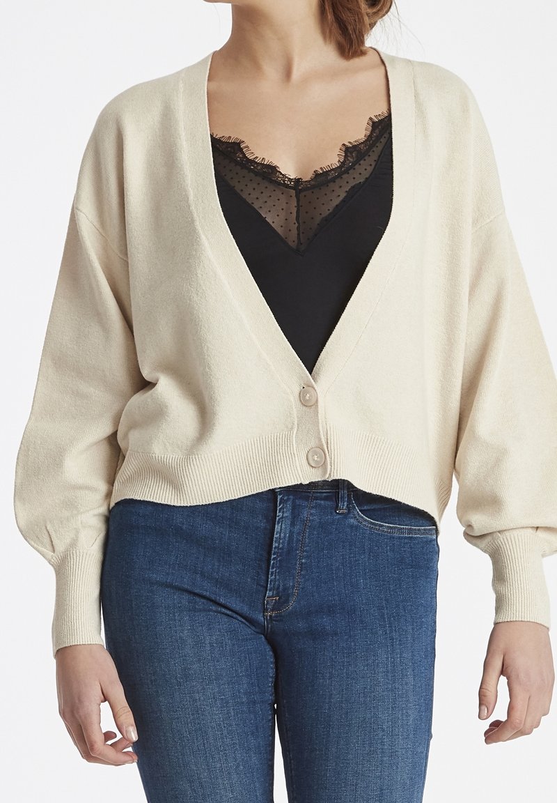 Vaaleanbeige neuleliivi, jossa on V-pääntie, lyhyt muoto ja kaksi etunappia. Siinä on leveät hihat ja joustinneule reunoilla. Käytetään mustan pitsitopin päällä.