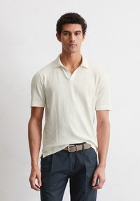 Short-sleeve, collared polo shirt in light cream, made of soft fabric. Paired with dark pants and a beige belt, featuring a simple design.