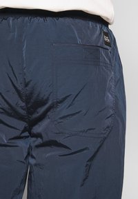 Närbild av marinblå glänsande byxor med elastisk midja, bakficka och liten svart etikett på högra sidan.