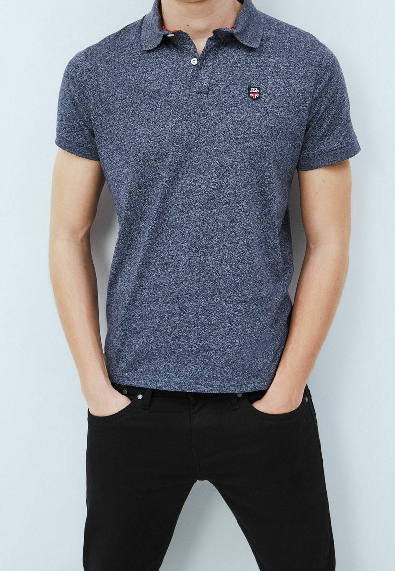 Homme portant un polo bleu marine à manches courtes bien ajusté avec un logo sur la poitrine et un pantalon noir, les mains dans les poches, sur un fond clair.