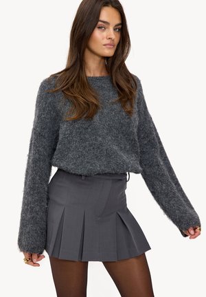 Pull gris duveteux à manches longues associé à une jupe plissée grise taille haute. Le mannequin porte des collants noirs transparents et une bague en or.