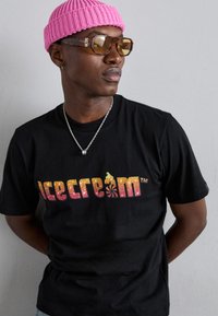 Sort T-shirt med et glitrende "is" logo i gradientfarver. Modellen bærer en lyserød strikket hue og brune solbriller samt en sølv kædedesign.