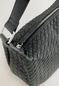 Schwarze gewebte Ledertasche mit rechteckiger Form, ausgestattet mit einem Reißverschluss und einem flachen Riemen. Strukturierte Oberfläche mit auffälligen Steppnähten.