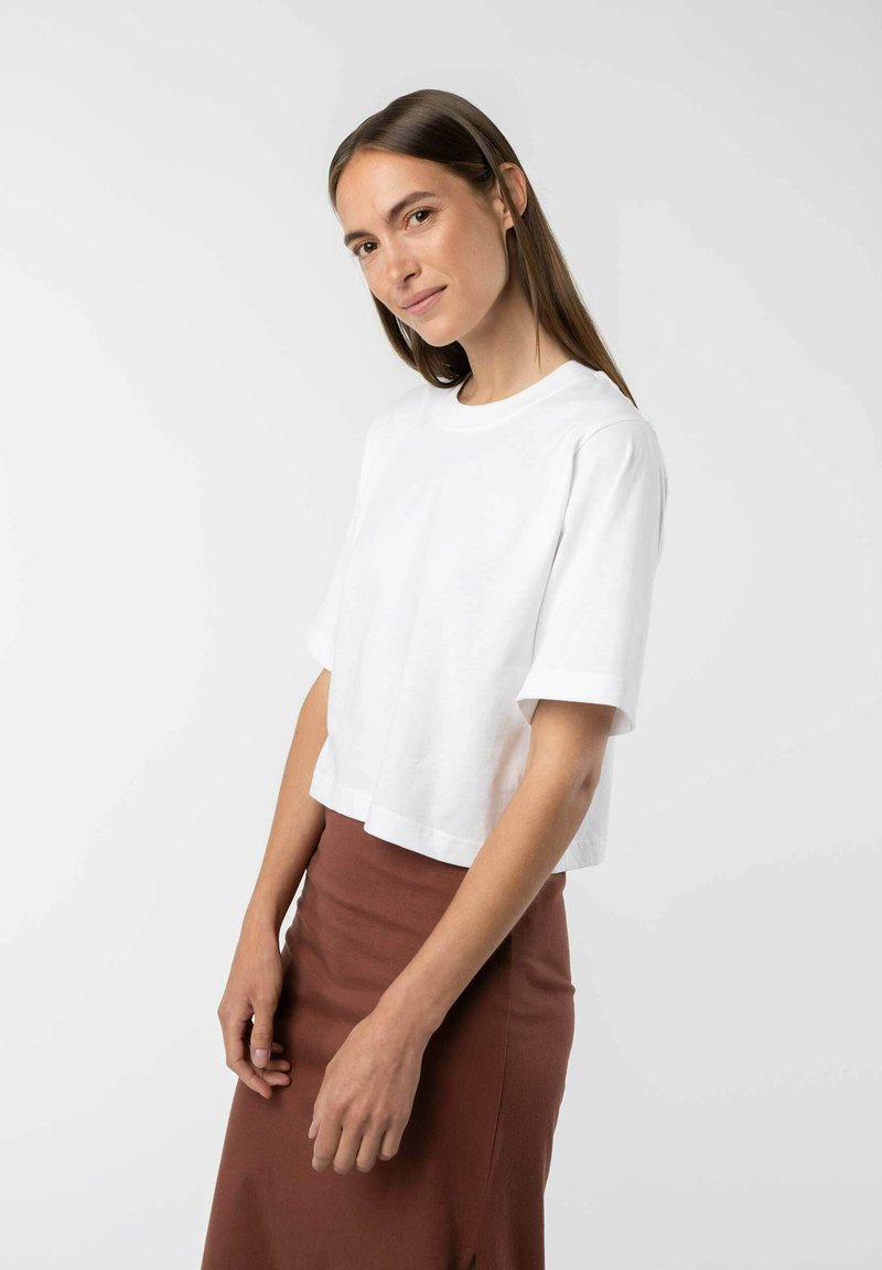Witte cropped t-shirt met korte mouwen, gemaakt van zacht materiaal. G gedragen met een bruine aansluitende rok. Eenvoudig, schoon ontwerp met een ronde halslijn.