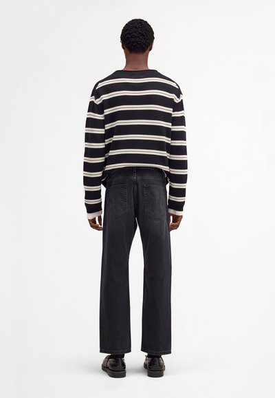 Henkilö, jolla on lyhyet kiharat hiukset ja joka pukeutuu mustavalkoraidalliseen neulepaitaan, mustiin leveälahkeisiin farkkuihin ja mustiin kenkiin, seisoo selin.