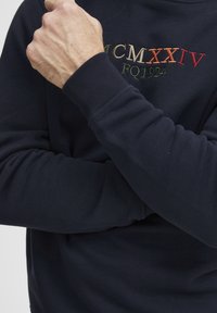 Sudadera azul marino con puños acanalados, que presenta texto bordado multicolor: "MCMXXIV FQ1924" con acentos en rojo, verde y naranja.
