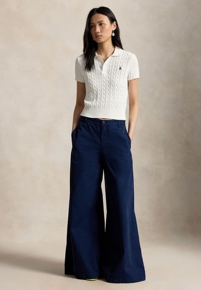 Woman standing wearing white short-sleeve cable knit polo and navy wide-leg pants, hands in pockets, against plain neutral background.