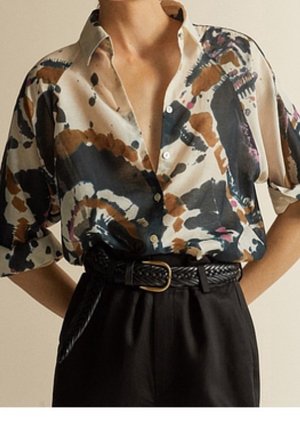 Kvinde iført en beige skjorte med knapper og sorte, brune og pink abstrakte mønstre, stukket ned i højtaljede sorte bukser med et fletbælte.