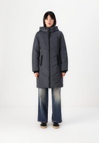 Manteau long en duvet gris foncé avec un motif de piquage en losanges, fermeture éclair à l'avant, capuche et poches latérales. Associé à un jean évasé et des baskets.