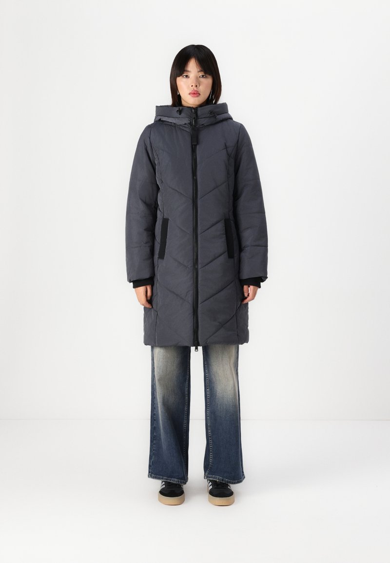 Manteau long en duvet gris foncé avec un motif de piquage en losanges, fermeture éclair à l'avant, capuche et poches latérales. Associé à un jean évasé et des baskets.