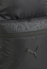 Mochila negra con una superficie suave y texturizada, adornada con patrones abstractos tonales sutiles. Incluye una cremallera negra y un acento de logo negro en relieve.