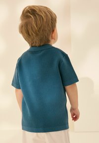Polo tričko s krátkým rukávem a vroubkovaným úpletem v modrozelené barvě, má límeček a volný střih, v kombinaci se světle zbarvenými kalhotami, z pohledu zezadu.