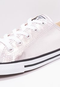 Metalické růžově zlaté tenisky s hladkým povrchem, bílými tkaničkami a odstínovým černobílým logem na jazyku. Gumová podrážka s černými pruhy.