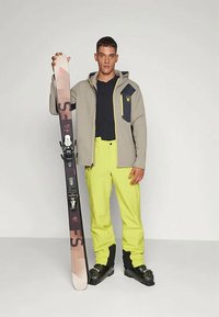 Homem com uma jaqueta cinza e calças de esqui amarelas brilhantes segurando um par de esquis na vertical, usando botas de esqui pretas, em fundo simples.