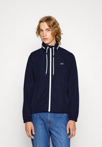 Veste coupe-vent marine avec fermeture éclair blanche et cordons de serrage, dotée d'un col haut et de poignets élastiques. Logo Lacoste sur la poche gauche de la poitrine.
