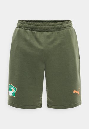 Olivzaļas sporta šorti ar elastīgu jostu, Kotdivuāras emblēma uz kreisās kājas un oranžs Puma logotips uz labās kājas.
