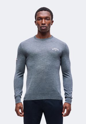 Homme portant un pull gris ajusté à manches longues avec un logo Callaway sur la poitrine, debout devant un fond blanc uni.