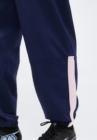 Πόδια που φορούν μπλε-σκούρο παντελόνι με ροζ ανοιχτή ρίγα στο πλάι και μαύρα αθλητικά παπούτσια με μπλε και λευκές λεπτομέρειες.