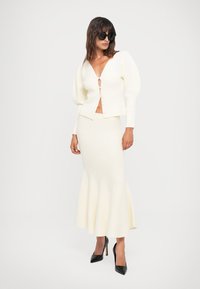 Bela rebrasta pletena kardigan z napihnjenimi rokavi, globokim V-izrezom in gumbom, v kombinaciji z usklajenimi razširjenimi krilom, obutimi v črne pete.