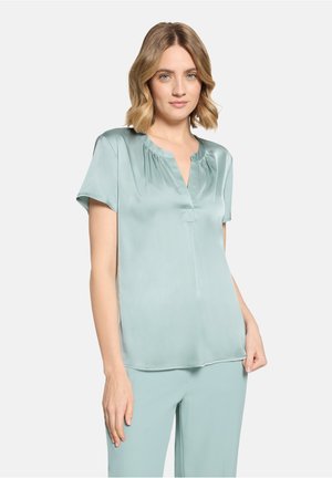 Jeune femme aux cheveux blonds mi-longs portant un chemisier bleu clair en satin à manches courtes et un pantalon assorti, debout devant un fond blanc.