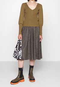 Olivgrüner gerippter Pullover, lange Ärmel, V-Ausschnitt; metallisch plissierter brauner Rock; Tasche mit schwarz-weißem Muster; olivgrüne Stiefel mit orangenen Sohlen.