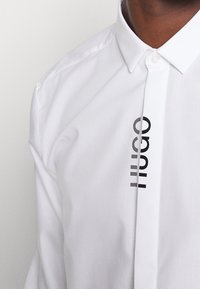 Witte shirt van gladde stof met een puntige kraag en een verticale zwarte logoprint aan de linkerkant.