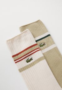 Dos pares de calcetines acanalados: uno crema con rayas rojas y beige, y otro verde oliva con rayas blancas y verdes. Ambos presentan un logo.