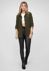 Groene ribfluwelen overhemdjas met knoopsluiting, opgerolde mouwen; gecombineerd met zwarte, hoog getailleerde legging en enkellaarzen.