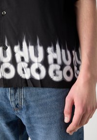 Chemise noire avec texte blanc en forme de flamme « HUGO » en bas, associée à un jean en denim bleu, montrant une main droite à côté de la hanche.