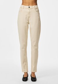 Pantaloni beige a taglio aderente indossati con ballerine nere, mostrati dalla vita ai piedi su uno sfondo chiaro uniforme.