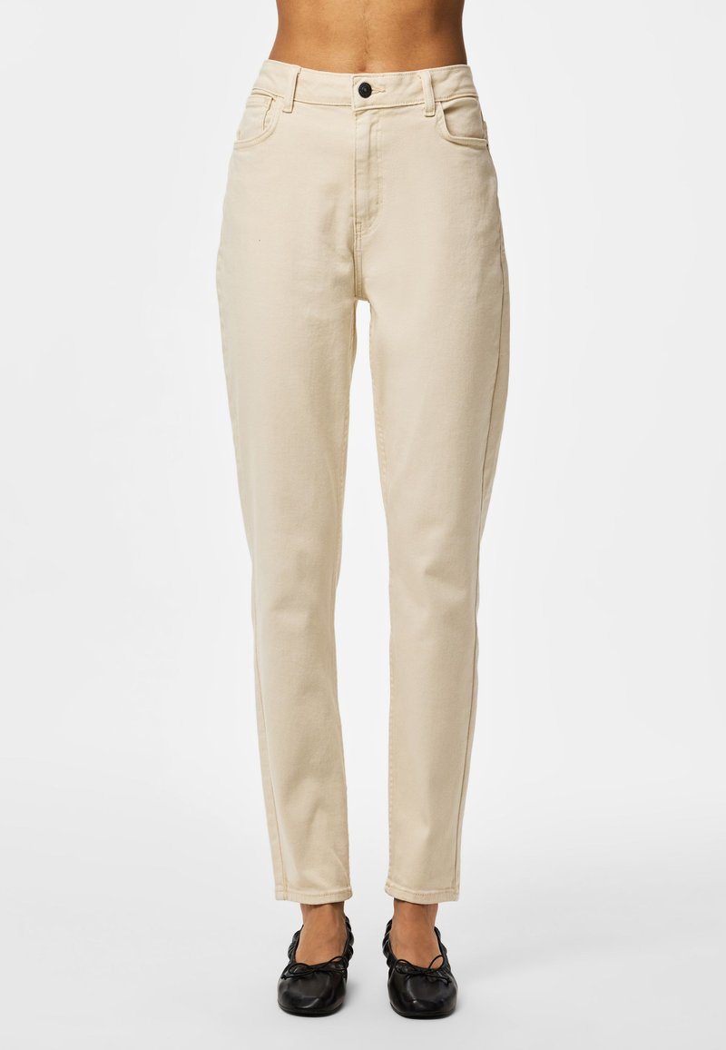Pantaloni beige a taglio aderente indossati con ballerine nere, mostrati dalla vita ai piedi su uno sfondo chiaro uniforme.