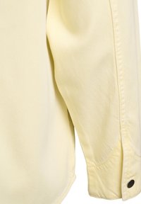 Detailné záber na svetložltý rukáv košele s čiernym cvokovým gombíkom na manžete, zobrazujúci textúru látky a detaily prešitia švov.