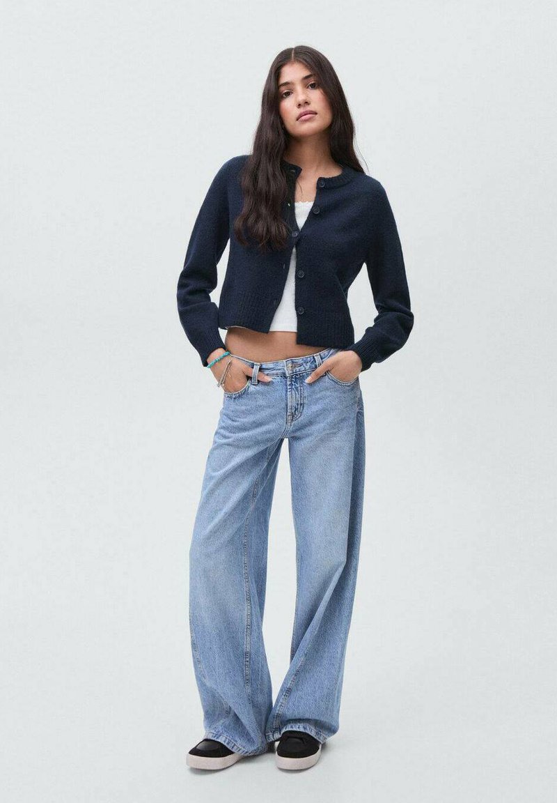 Ung kvinna står med händerna i fickorna, klädd i en marinblå kofta, vit crop-top, vida ljusblå jeans och svarta sneakers.