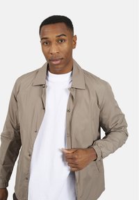 Lekka beżowa kurtka z kołnierzykiem, zapięciem na napy z przodu i elastycznymi mankietami. Nosi się na białym t-shircie. Gładka struktura.