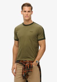 Camiseta de cuello corto en verde oliva con acentos en verde oscuro, con un pequeño logo en el pecho. Llevada con pantalones beiges y una camisa de cuadros atada a la cintura.