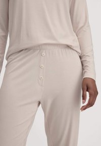 Nahaufnahme einer Person in einem hellbeigen Langarmoberteil und passenden Hosen mit einem elastischen Bund und drei vorderen Knöpfen.
