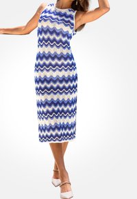 Mujer vestida con un vestido sin mangas, hasta la rodilla, con un patrón ondulado azul y blanco, y tacones blancos con correas de cadena dorada.
