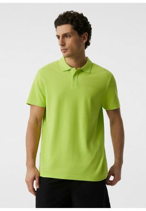 Mężczyzna z krótkimi kręconymi włosami, ubrany w jasnokrótki zielony podkoszulek polo i czarne szorty, patrzący w lewo na jednolity jasny tło.