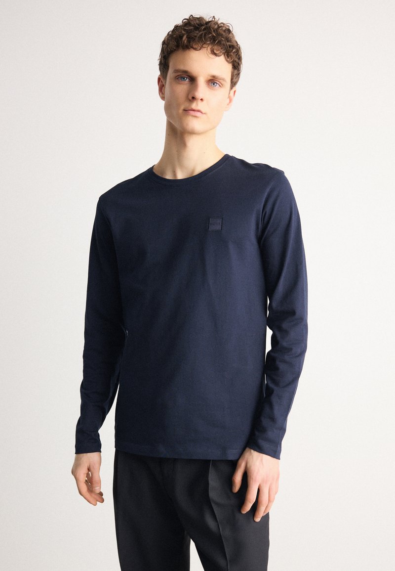 Camisa de manga larga de color marino, confeccionada en algodón, con un escote redondo y un pequeño parche negro con logo en el pecho. Textura suave.