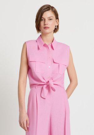 Femme aux cheveux bruns courts portant une chemise sans manches rose boutonnée nouée à la taille et un pantalon taille haute rose assorti, debout devant un fond blanc.