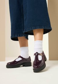 Zapatos Mary Jane brillantes de color púrpura con un diseño de tira en T, suela negra gruesa y hebilla de acento, usados con calcetines blancos hasta el tobillo y amplios culottes de mezclilla.