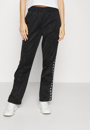 Mujer vistiendo pantalones deportivos negros con rayas blancas con el logo a los lados y zapatillas blancas, de pie contra un fondo blanco liso.