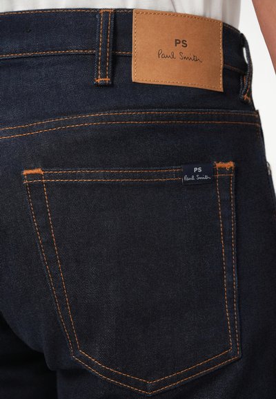 Jeans de ganga escura com costura laranja em contraste, apresentando uma etiqueta de couro rotulada "PS Paul Smith" na cintura e uma pequena etiqueta da marca no bolso.