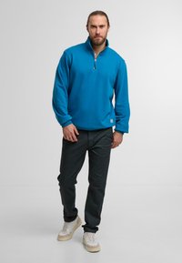 Turkusowy polarowy sweter z ćwierć zamkiem, prążkowanymi mankietami i miękką teksturą, zestawiony z ciemnoszarymi spodniami i białymi sneakersami.