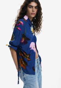 Floral button-up shirt in blue, featuring large multicolored flowers. Soft fabric, with adjustable tie details on the sides and a classic collar.